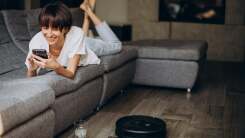 woman lying on couch, using her phone, while a robot vacuum cleans the floor