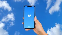 A hand holding a smartphone displaying the BlueSky logo, on a background of a blur cloudy sky