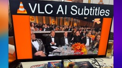 A TV screen that displays the cone logo of VLC media player, followed by the text VLC AI Subtitles. The TV is paused at one frame in a scene, with subtitles displayed below the video.