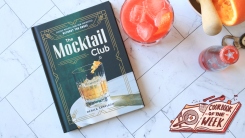 A mocktail cookbook on a counter with juiced oranges and a red drink.