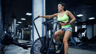 A woman in Spandex exercide gear works on an elliptical machine in an otherwise empty gym filled with workout equipment.