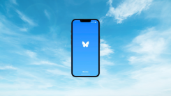 A smartphone showing the Bluesky app loading screen superimposed on a background of a bright blue sky