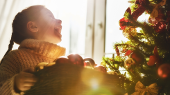 A young girl laughing as she decorates an indoor Christmas tree with ornaments taken from a box
