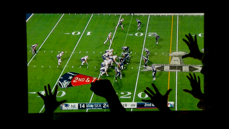 The Super Bowl playing on a flatscreen TV in a dark room with viewers visible cheering in front of it
