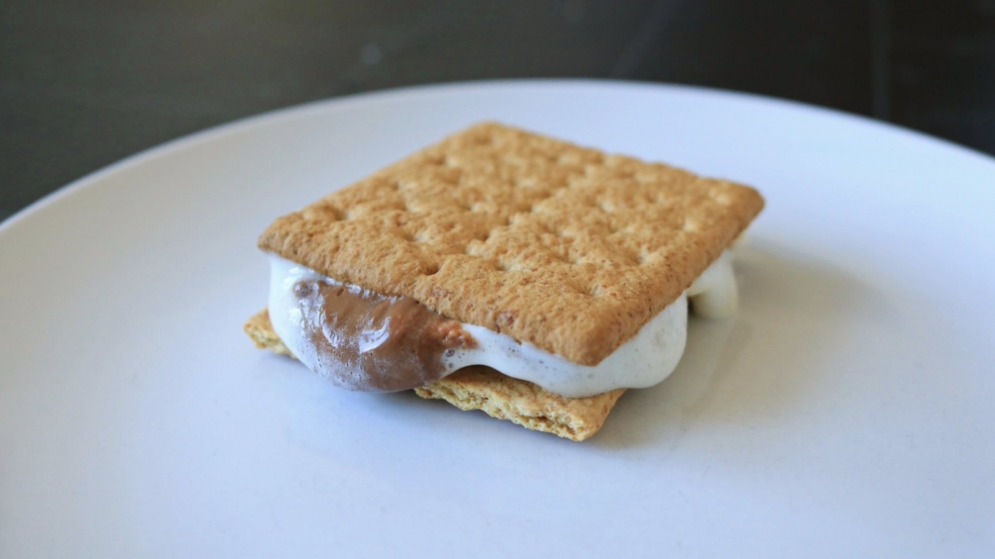 A s'more with melted chocolate and marshmallow oozing out on a plate.